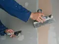 Ręce w niebieskim kombinezonie nakładają gładź polimerową na ścianę za pomocą szpachelki, przygotowując ją do malowania.