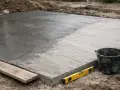 Świeżo wylany beton na placu budowy. Poziomica i wiadro obok. Czas wiązania betonu B25 zależy od warunków.