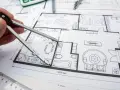 Rysunek techniczny mieszkania, cyrkiel w dłoni. Kierownik budowy analizuje projekt, rozważając jakie zmiany w projekcie może wprowadzić, by usprawnić budowę.