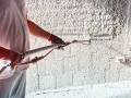 Ręce w rękawiczkach trzymają pistolet do nakładania tynku na ścianę. Powierzchnia jest pokryta grubą warstwą białego materiału.