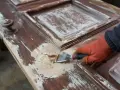 Ręka w pomarańczowej rękawicy zeskrobuje starą farbę z drewnianych drzwi.