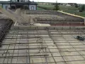 Budowa monolitycznego stropu zbrojonego. Drewniane deski i stalowe pręty tworzą siatkę gotową na zalanie betonem. W tle domy i zielone pola.
