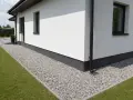 Biały dom z czarną rynną i żwirową opaską wokół domu, otoczony zielonym trawnikiem.