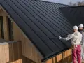 Dwóch budowlańców w kaskach pracuje na czarnym, metalowym dachu o nachyleniu 35 stopni. Drewniana elewacja domu kontrastuje z nowoczesnym pokryciem.