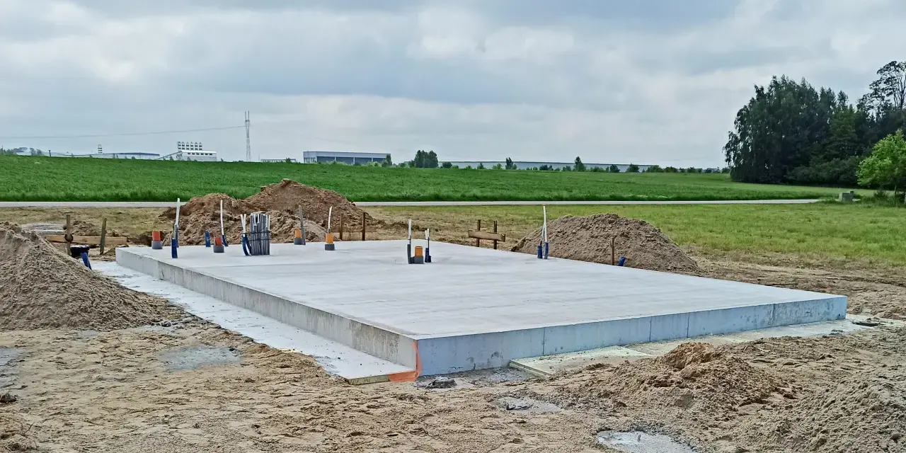 Płyta betonowa, czyli fundament, gotowa do budowy domu. Wokół piasek i zielone pola.
