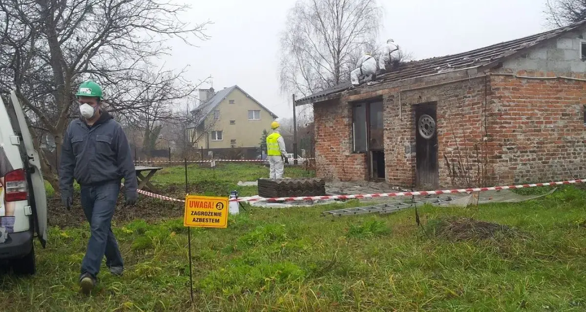 Pracownicy w kombinezonach usuwają eternit z dachu. Tablica ostrzega przed zagrożeniem azbestem.
