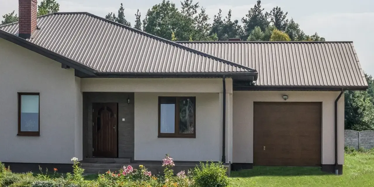 Dom z dachem z blachy trapezowej. Wymiary dachu dopasowane do nowoczesnej bryły budynku. Przed domem ogród z kwiatami.