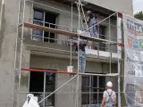 Pracownicy na rusztowaniu warszawskim montują materiały. Widać fragment budynku z oknami i baner reklamowy.