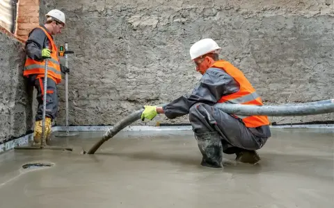 Dwóch budowlańców w kamizelkach odblaskowych wylewa pianobeton. Jeden kieruje wężem strumień materiału, drugi kontroluje poziom.