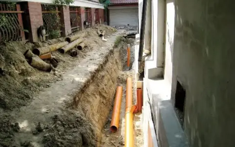Wykop pod drenaż wokół starego domu, widoczne nowe pomarańczowe rury i stare, zniszczone rury.