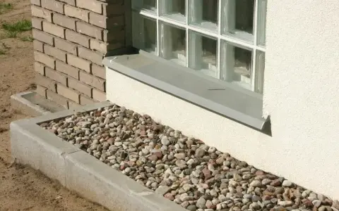 Kamienna opaska żwirowa wokół domu, otaczająca betonową rabatę z kolorowych kamieni, stanowi estetyczne i praktyczne wykończenie.
