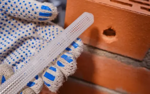 Ręka w rękawicy trzyma biały, siatkowy kołek do kotwy chemicznej obok cegły z otworem.