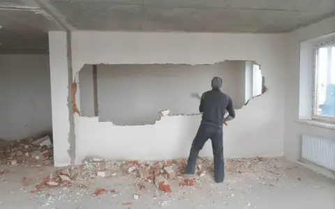 Mężczyzna z młotem wyburza ścianę nośną, tworząc w niej duży otwór. Wokół leżą gruzy.