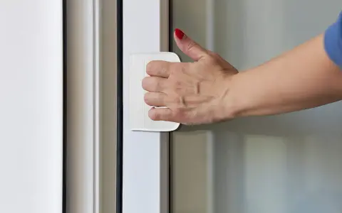 Dłoń z czerwonym lakierem na paznokciach naciska na biały zatrzask drzwi balkonowych.
