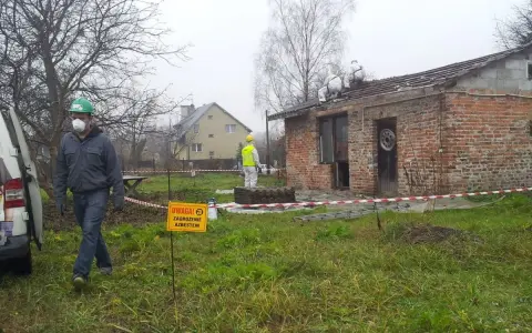 Pracownicy w kombinezonach usuwają eternit z dachu. Tablica ostrzega przed zagrożeniem azbestem.