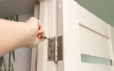 Dłoń dokręca śrubę w zawiasie drzwi, wykonując regulację zawiasów drzwi.