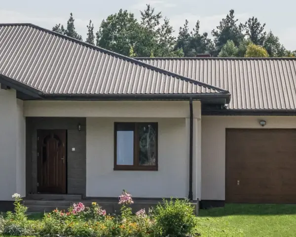 Dom z dachem z blachy trapezowej. Wymiary dachu dopasowane do nowoczesnej bryły budynku. Przed domem ogród z kwiatami.