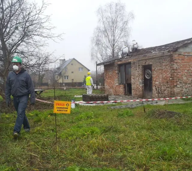 Pracownicy w kombinezonach usuwają eternit z dachu. Tablica ostrzega przed zagrożeniem azbestem.