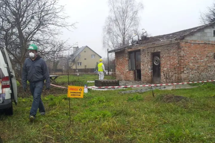 Pracownicy w kombinezonach usuwają eternit z dachu. Tablica ostrzega przed zagrożeniem azbestem.