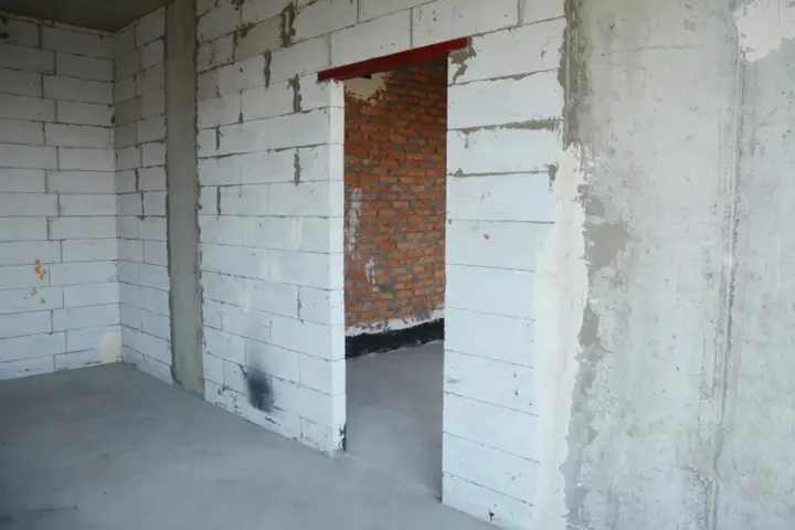 Wnętrze w budowie. Widoczny otwór drzwiowy z czerwonym nadprożem stalowym, ściany z białych pustaków i cegły.