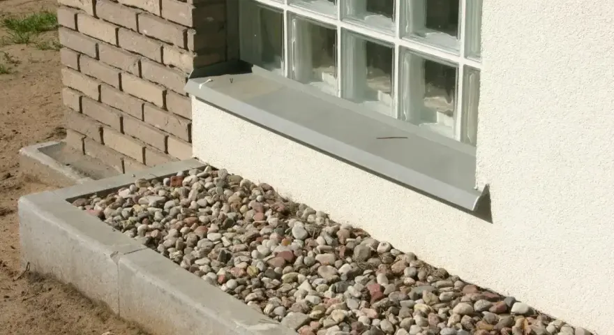 Kamienna opaska żwirowa wokół domu, otaczająca betonową rabatę z kolorowych kamieni, stanowi estetyczne i praktyczne wykończenie.