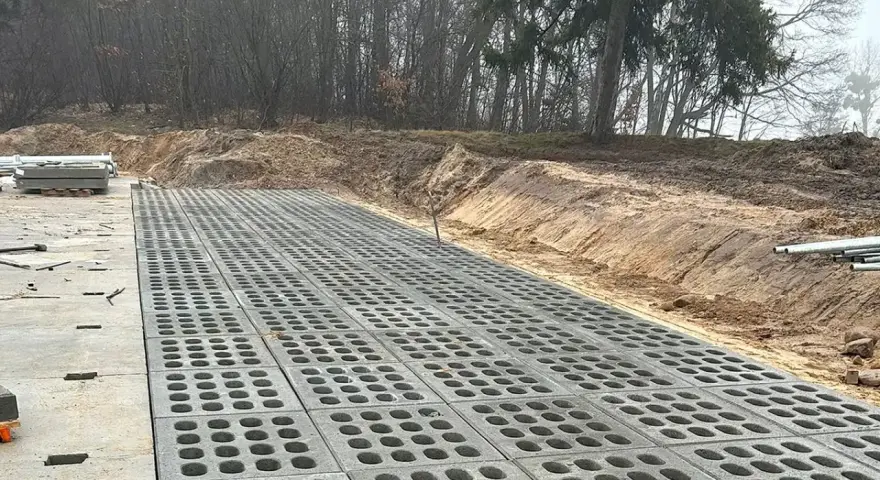Trwa utwardzenie terenu. Betonowe płyty z otworami tworzą nawierzchnię obok placu budowy z materiałami.