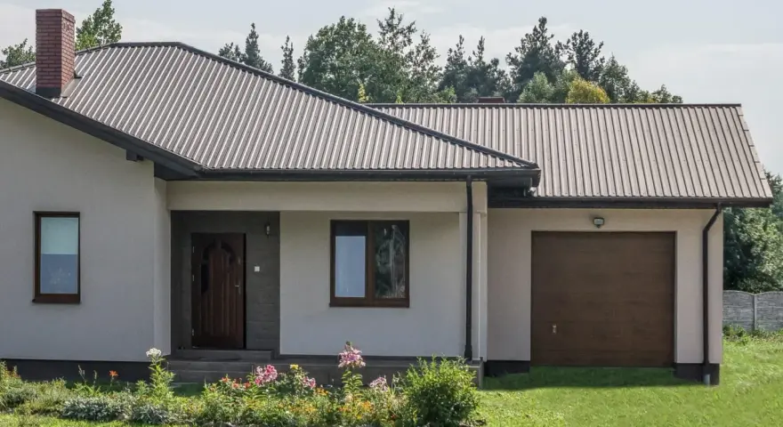 Dom z dachem z blachy trapezowej. Wymiary dachu dopasowane do nowoczesnej bryły budynku. Przed domem ogród z kwiatami.
