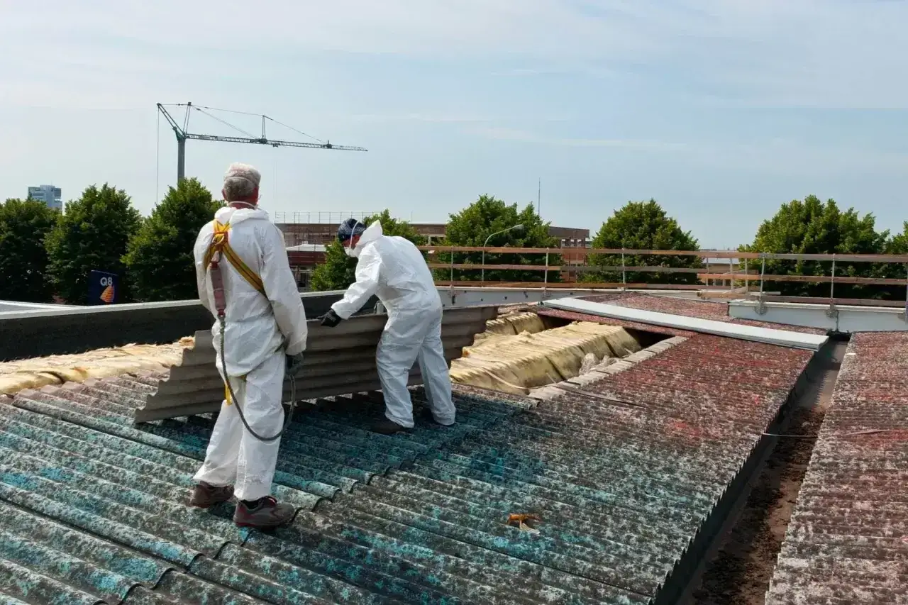Dwóch pracowników w kombinezonach ochronnych usuwa z dachu eternit, materiał zawierający azbest.