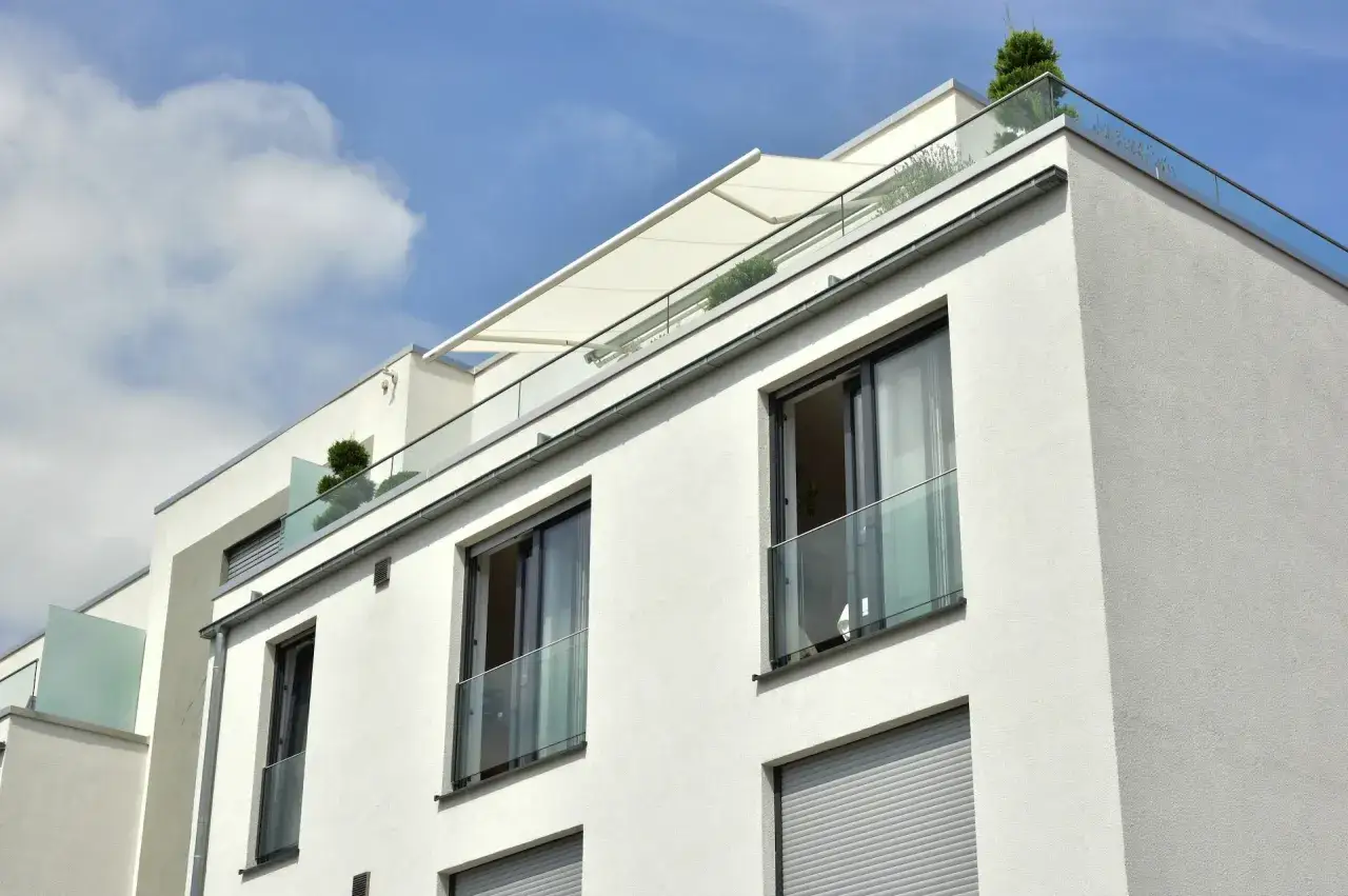 Nowoczesny budynek z białym tynkiem, szklanymi balustradami i rozkładanym markizą. Widoczne okno francuskie z widokiem na niebo.