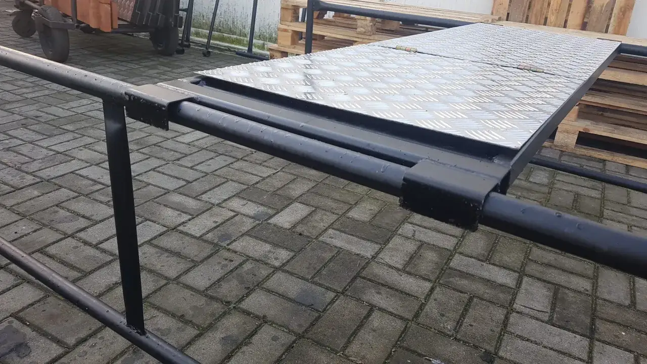 Metalowa platforma rusztowania warszawskiego, z widocznymi wymiarami i antypoślizgową powierzchnią, na tle palet i kostki brukowej.