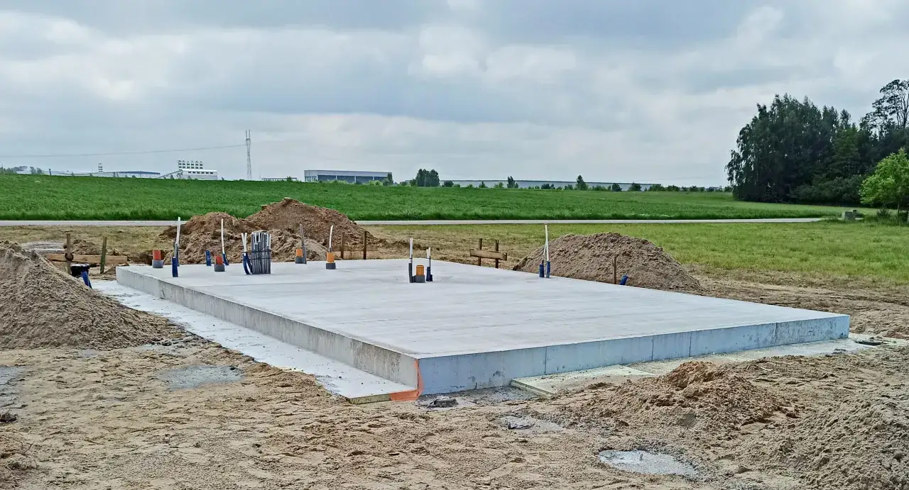 Solidny fundament, czyli płyta betonowa, czeka na budowę domu. Wokół piasek i zielone pola.