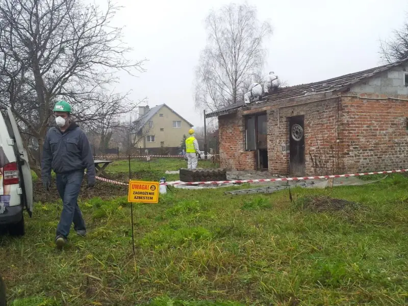 Pracownicy w kombinezonach usuwają eternit z dachu. Tablica ostrzega przed zagrożeniem azbestem.